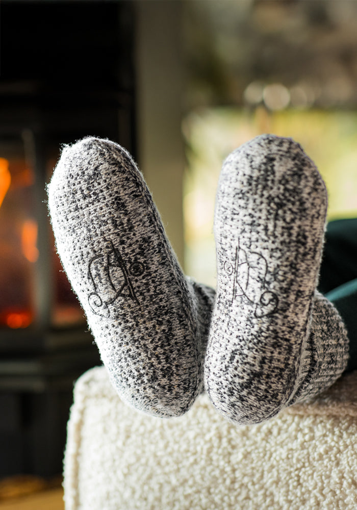 A close-up of a person wearing Woolx Women's Gripper Slipper Booties in Speckled Stone, resting their feet on an ottoman near a cozy fireplace.