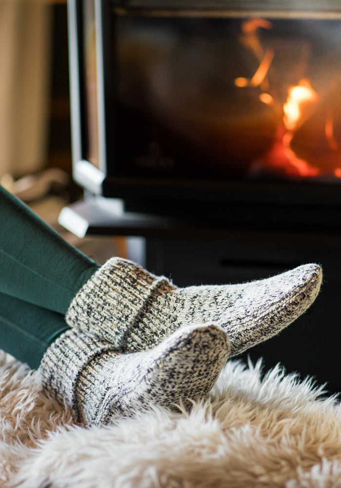 A person relaxes by the fire, feet snug in Woolx Women's Gripper Slipper Booties – Speckled Stone, resting on a plush rug.
