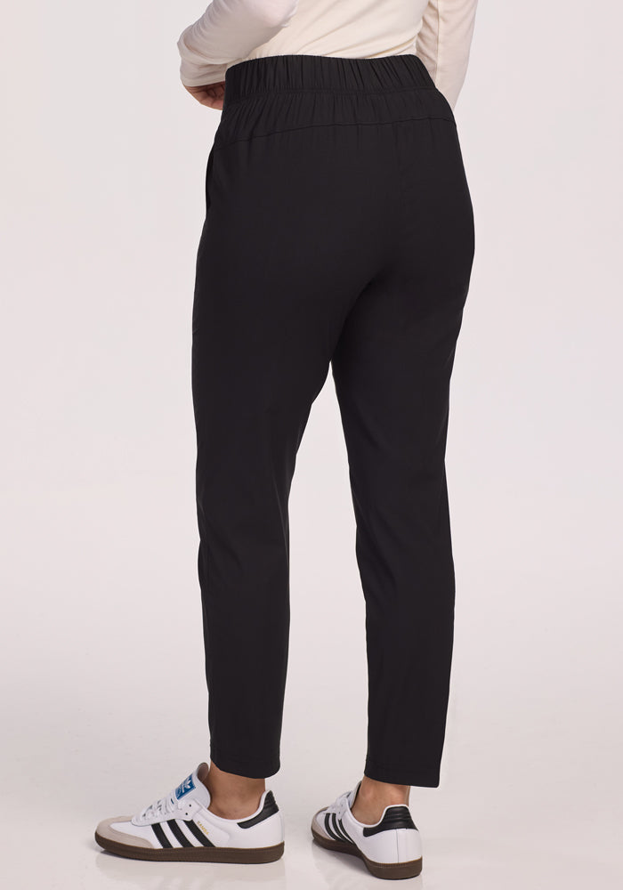 A person is shown from the back against a plain light background wearing Woolx Nicole Pants in black, paired with a light long-sleeve top and white sneakers with black stripes.