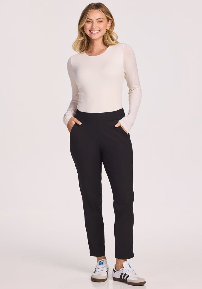 A woman smiles with her hands in the pockets of her Woolx Nicole Petite Ankle Pant. She pairs the pants with a long-sleeve white top and white sneakers with black accents, standing against a plain, light background.