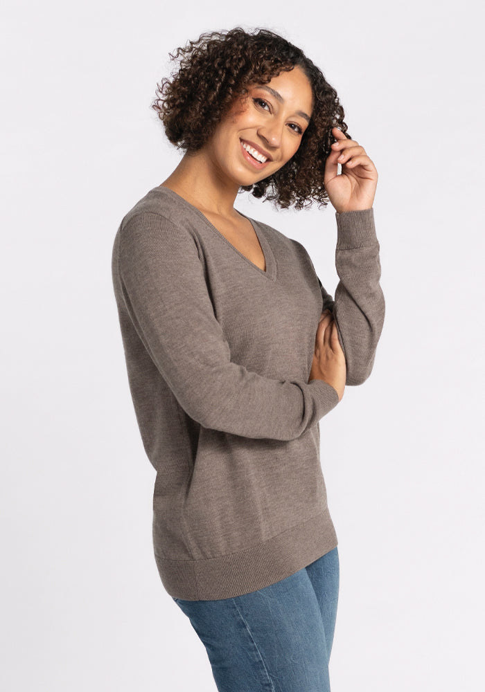 A woman with curly hair wears the Woolx Francesca Sweater in sable, paired with blue jeans, and smiles while posing with a hand in her hair, highlighting its luxurious comfort against a plain white background. Final Sale Deals!.