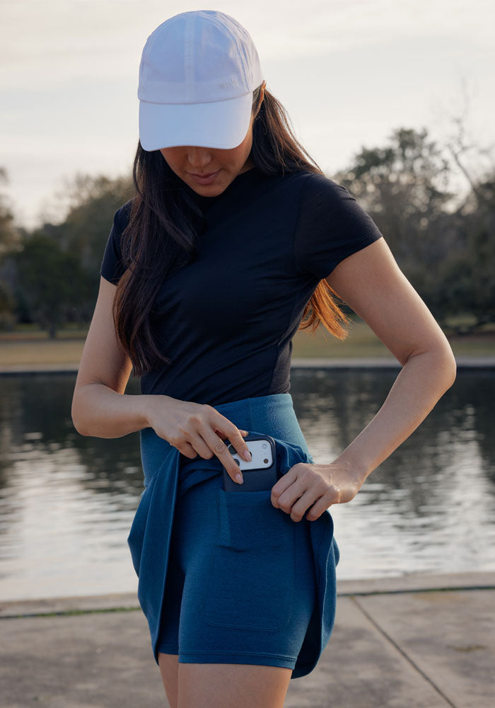 A woman in a white cap and black shirt stands by a lake, slipping her smartphone into the pocket of her Woolx Sienna Skort in Majolica Blue, with trees and water visible in the background.