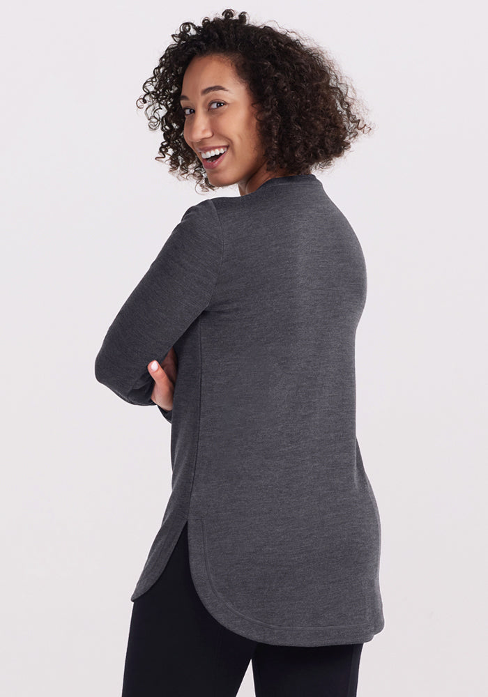 A woman with curly hair smiles over her shoulder, wearing the Woolx Blair Tunic in Cinder Melange with black pants, against a light background—embracing effortless style and lightweight Merino wool warmth.