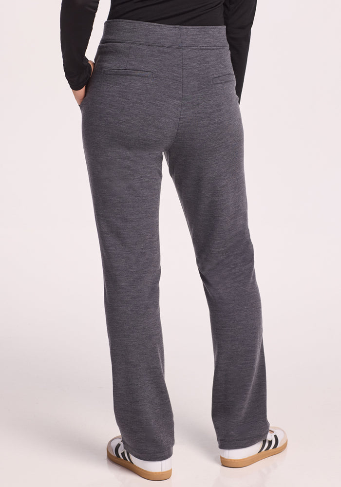 A person wearing Woolx Emerson Straight Leg Pants in Cinder Melange, a black long-sleeve top, and white sneakers with black stripes stands with hands in back pockets, facing away from the camera against a light backdrop.