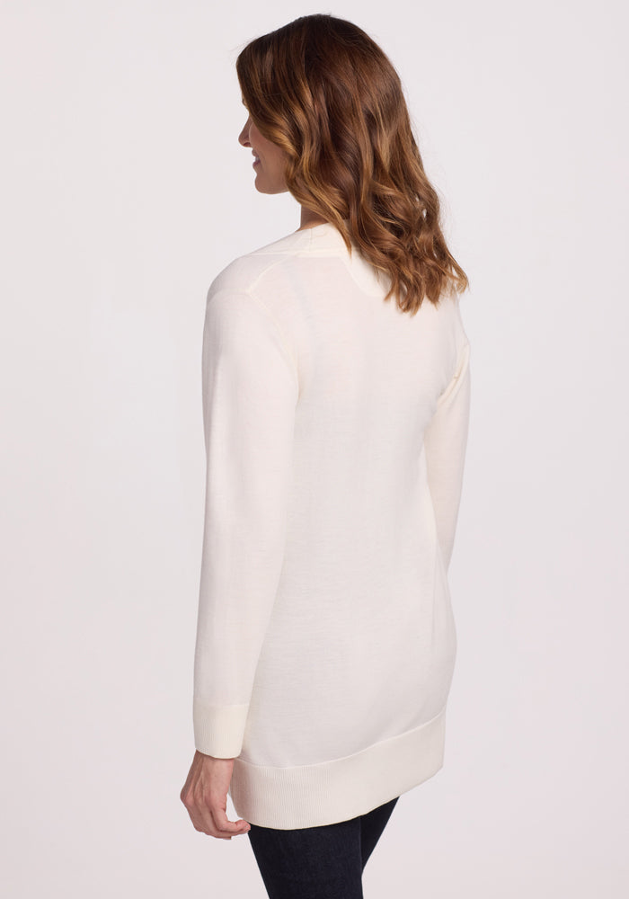 A woman with wavy brown hair stands with her back to the camera, wearing the Woolx Macie Sweater in Dewdrop Pearl and dark pants against a plain light background.