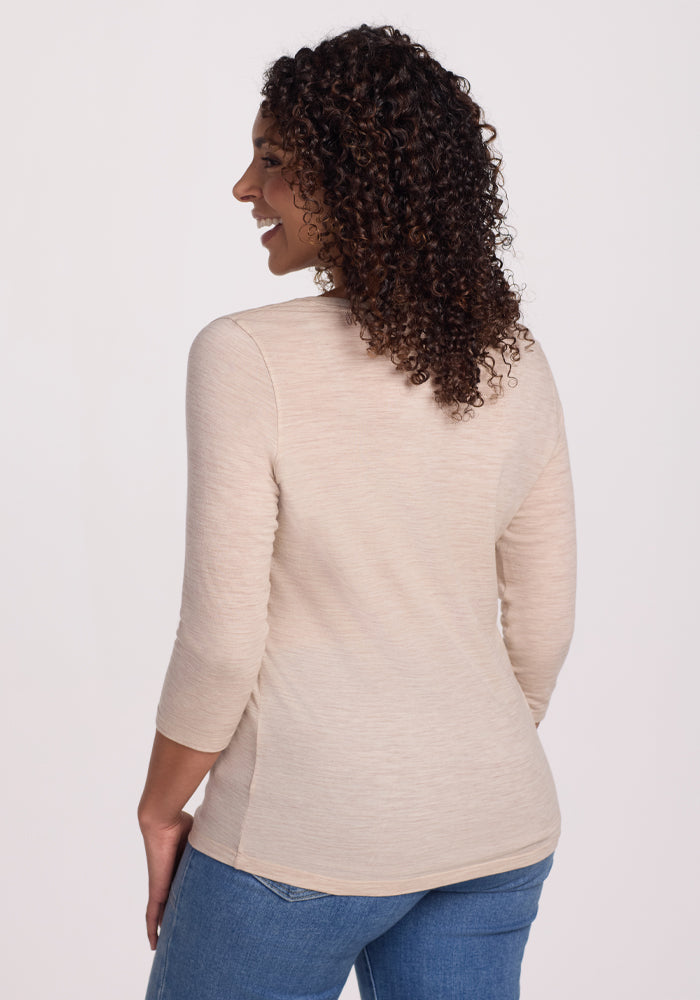 A person with curly hair stands smiling over their shoulder wearing a casual Jenny 3/4 Sleeve Crew Neck in Cream Heather by Woolx and blue jeans, set against a plain white background.