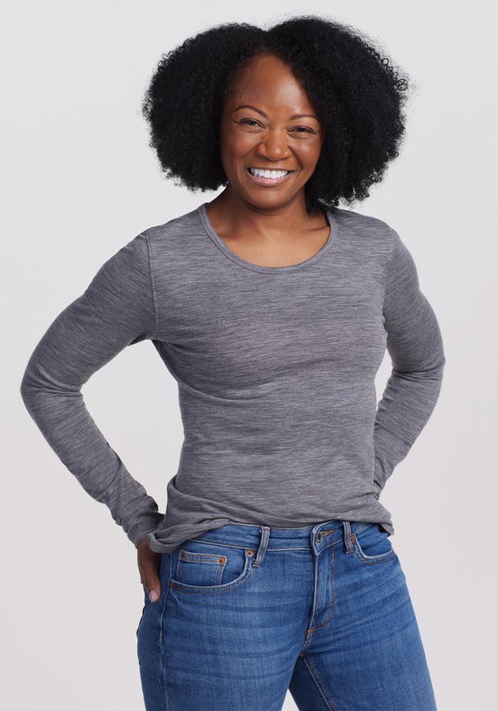 A curly-haired person in a Woolx Remi Long Sleeve T-Shirt - Graphite Heather and blue jeans smiles with hands on hips against a plain white background. 