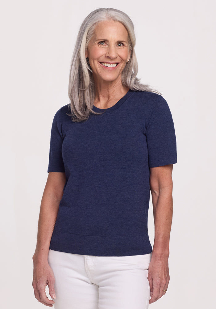A smiling older woman with long gray hair wears the Woolx Anna Crew Sweater in Twilight Storm and white pants, standing against a plain light background.