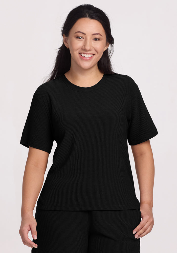 A person with long dark hair smiles, wearing the luxurious Woolx Delia Ribbed Top in black, paired with matching pants. The temperature-regulating outfit stands out against the plain, light-colored background. 