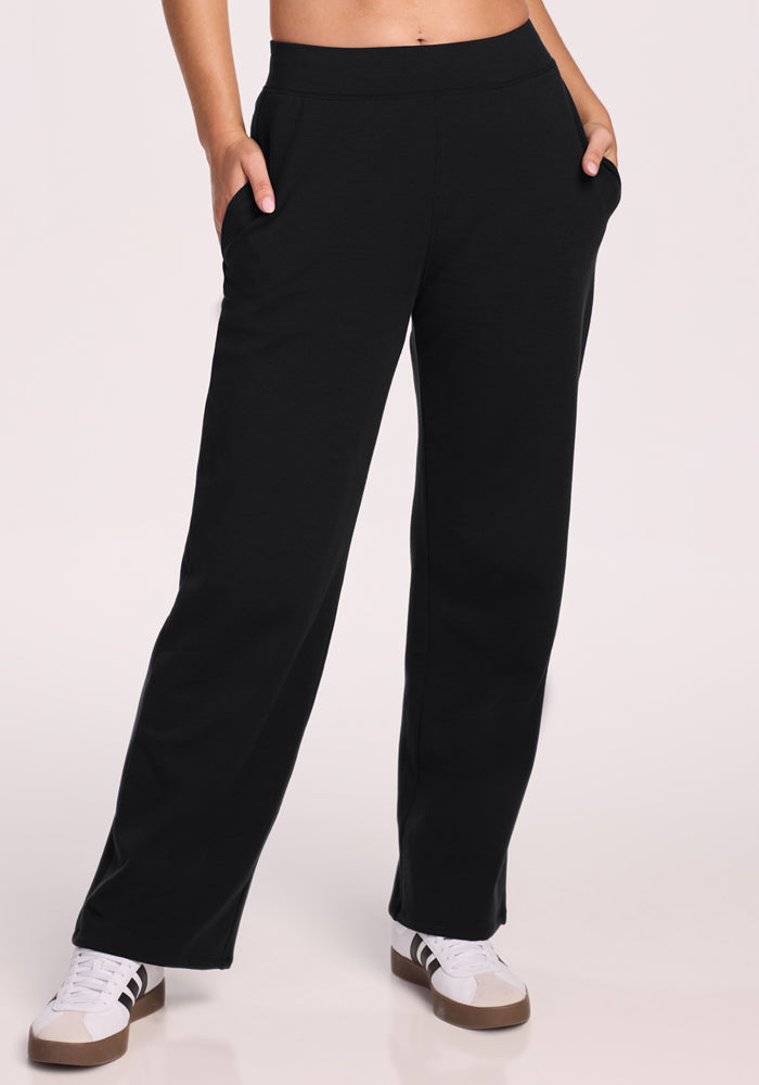 A person wearing Woolx Brynn Petite Pants in black, hands in pockets, paired with white sneakers featuring brown soles, stands against a plain light background.
