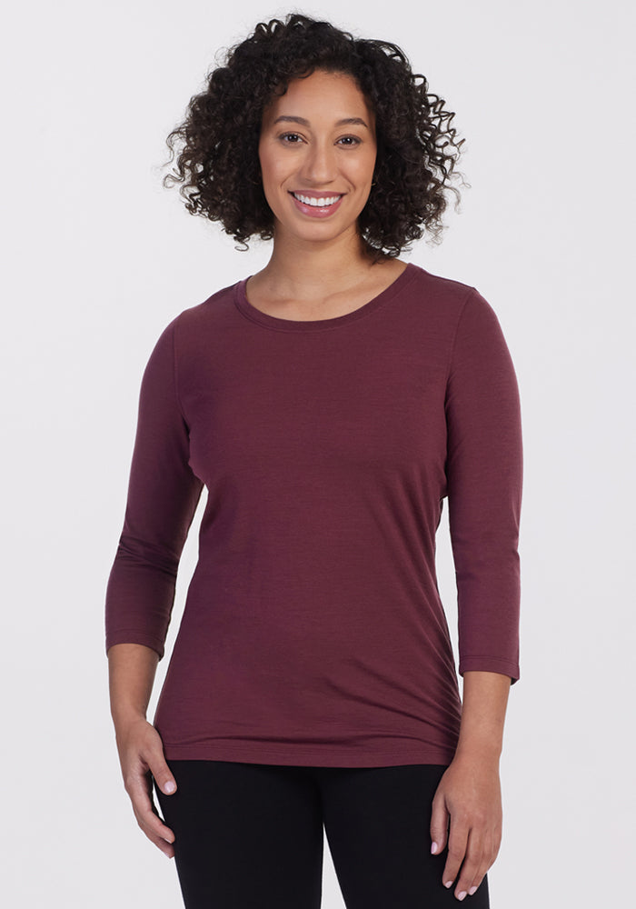 A woman with curly hair smiles against a white background, wearing Woolx’s Jenny 3/4 Sleeve Crew Neck in Grape Harvest and black pants.