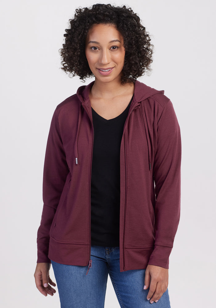 A person with curly hair faces the camera wearing the Woolx Ryann Hoodie • Final Sale - Grape Harvest over a black top and blue jeans, set against a plain, light background.
