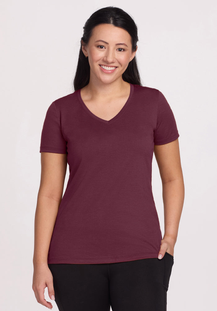 A woman with long dark hair smiles wearing the Woolx Junie V Neck in Grape Harvest and black pants, standing against a plain light background.