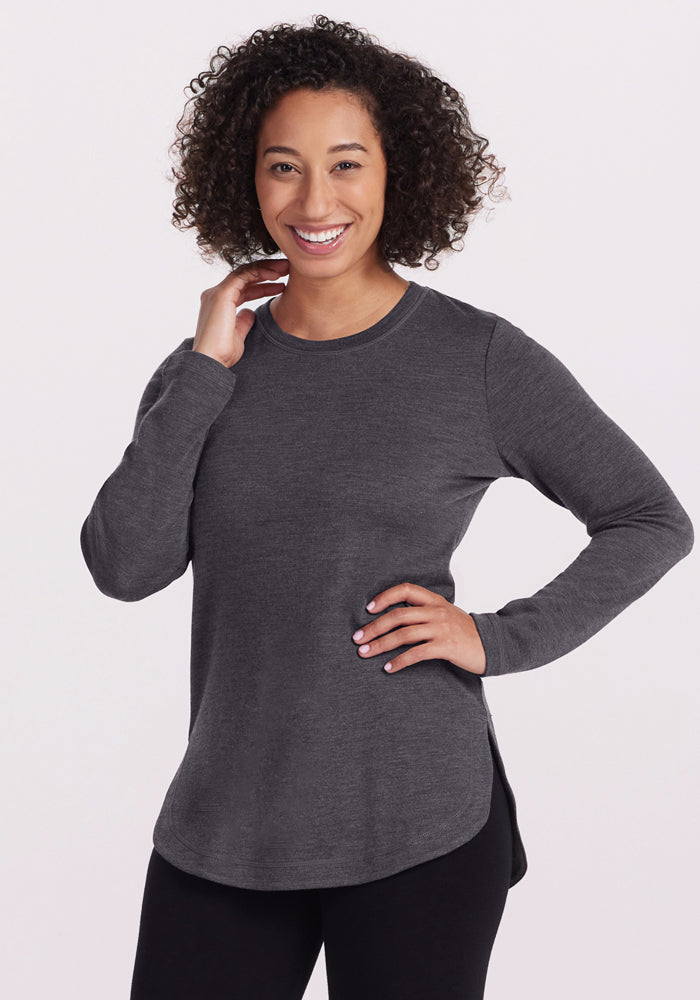 A woman with curly hair smiles at the camera in the Woolx Blair Tunic - Cinder Melange, paired with black pants. She rests one hand on her hip and touches her neck, highlighting the tunic’s lightweight warmth against a plain light background. 
