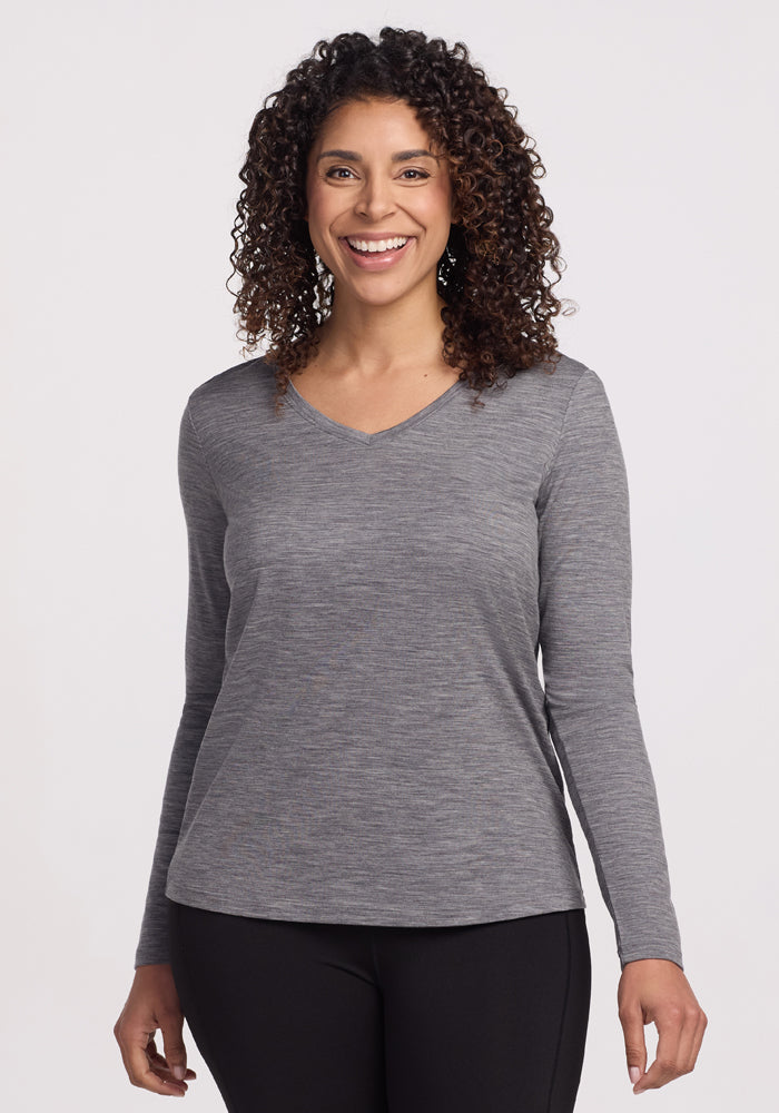 A person with curly hair is smiling in a Woolx Millie Long Sleeve in Graphite Heather and black pants, standing against a plain white background. 