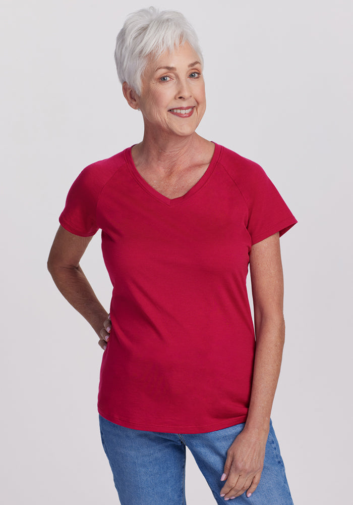 Smiling older woman in a Woolx Mia V Neck (Poppy Spark) shirt and blue jeans, standing against a plain background.