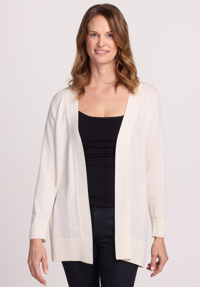A woman with shoulder-length brown hair wears the Woolx Macie Sweater in Dewdrop Pearl over a black top and dark jeans. Against a plain white background, she smiles slightly at the camera. 