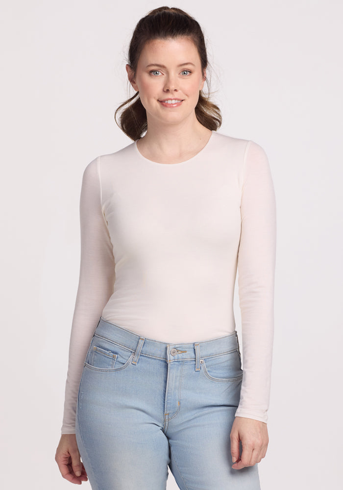 A person with long hair wearing a Woolx Sophia Bodysuit in Vanilla and light blue jeans stands smiling gently at the camera against a plain background. 