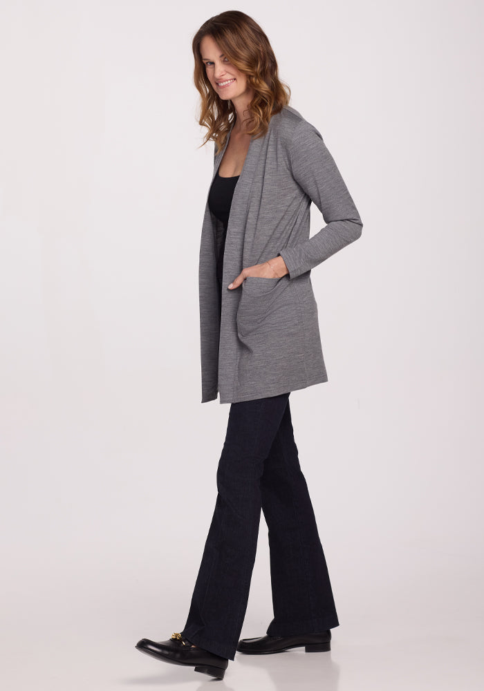 A smiling person with long, wavy hair walks left, wearing dark pants and black shoes. They're wrapped in Woolx's feather-light Paisley Cardigan in Graphite Heather over a black top. The plain white background emphasizes their comfort.