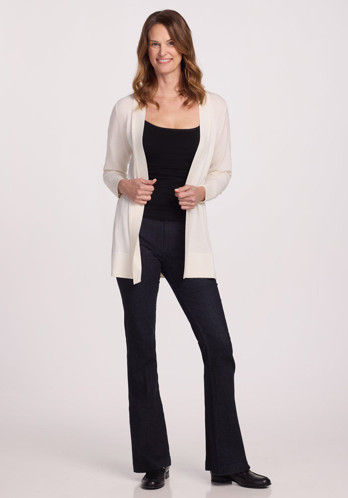 A person with long hair stands smiling in a Woolx Macie Sweater - Dewdrop Pearl over a black top and dark pants, with one hand in their pocket and wearing black shoes. The background is plain white.
