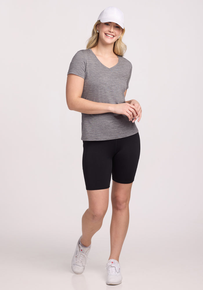 A smiling woman stands mid-step against a plain background, wearing the Woolx Junie V Neck in Graphite Heather with black bike shorts, white sneakers, and a white cap. She looks casual and relaxed with one hand on her hip.