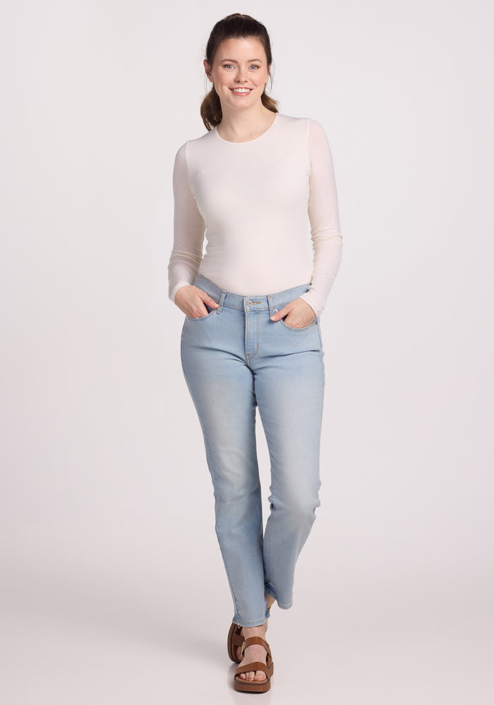 A person with long hair tied back is wearing a vanilla Sophia Bodysuit from Woolx and light blue jeans, smiling at the camera. Their hands are in their pockets as they stand against a plain white background, sporting brown sandals.
