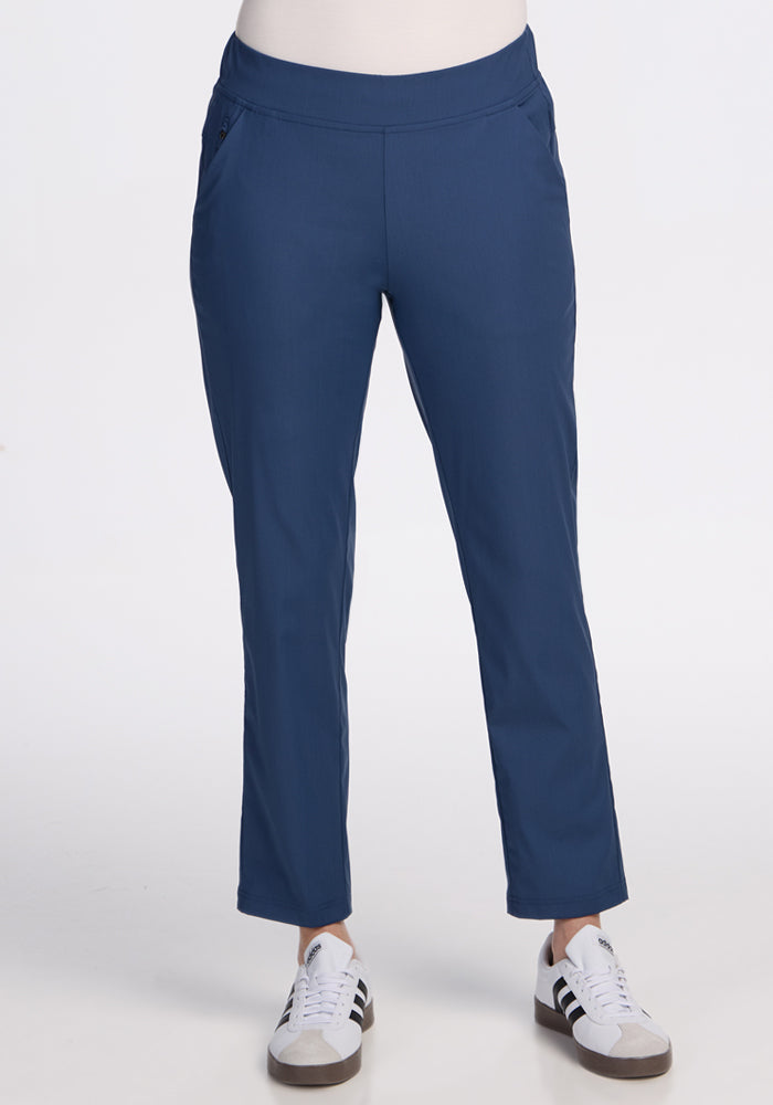 A person wearing Woolx Nicole Ankle Pants in Vintage Indigo pairs them with blue and white sneakers featuring black stripes, standing against a plain white background with hands in pockets. Only the lower half of their body is shown. 