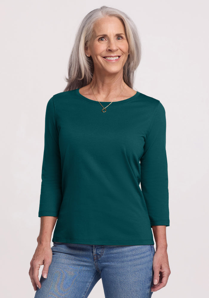 Smiling older woman with long gray hair wearing the Woolx Jenny 3/4 Sleeve Crew Neck in Riverbank Green and blue jeans, standing against a light background.