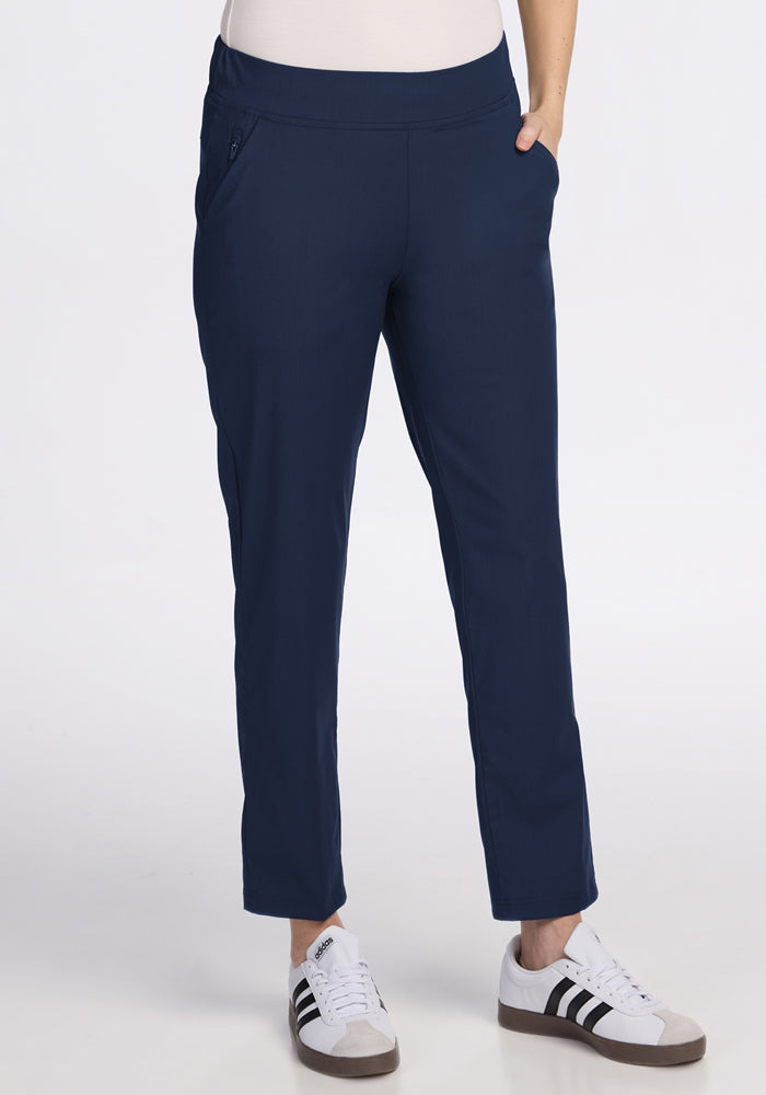A person wearing Woolx Nicole Petite Ankle Pant in Deep Navy with hands in pockets and white sneakers with black stripes, standing against a plain white background.
