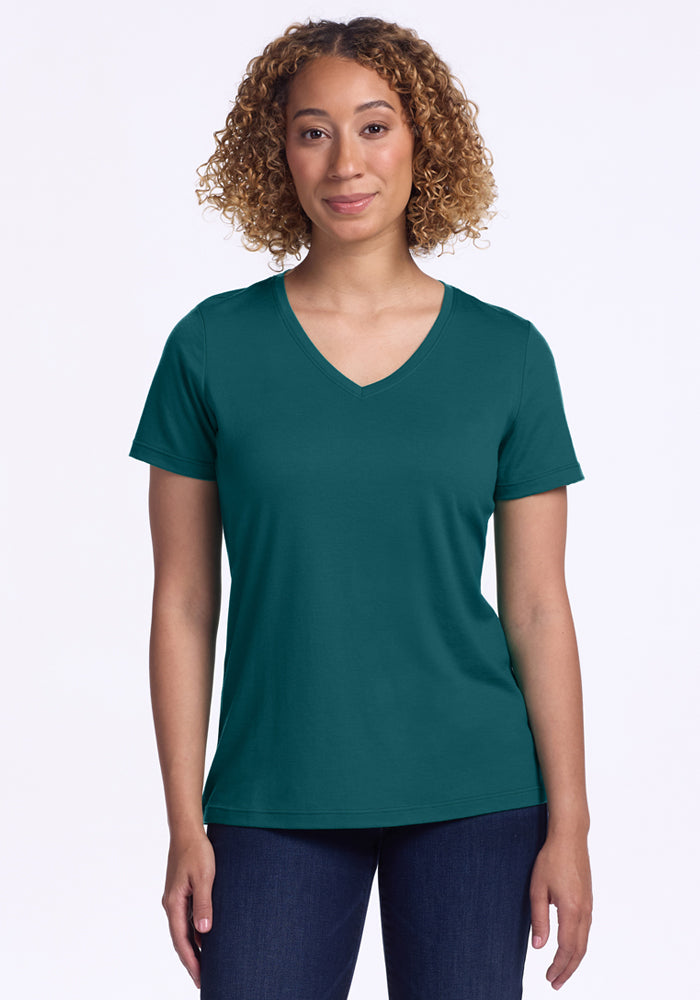 A woman with curly hair wears the Woolx Junie V Neck in Riverbank Green and dark jeans, standing against a plain white background and smiling softly at the camera.
