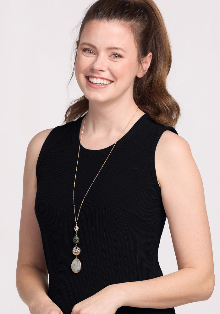 A person with long brown hair tied back smiles in a Woolx Iris Ribbed Dress - Black, paired with a long pendant necklace against a plain light-colored background.