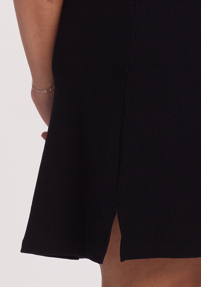 The person is wearing the Harlowe Ribbed Dress by Woolx in black, featuring a slit that reveals part of the leg. A delicate silver bracelet graces the wrist.