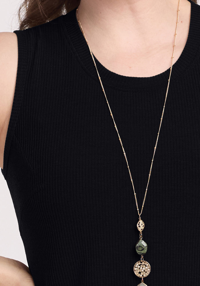 A model wearing the Iris Ribbed Dress in black by Woolx pairs it with a long, delicate gold necklace featuring two intricately designed pendants. The image highlights the dress's upper half and accessories.