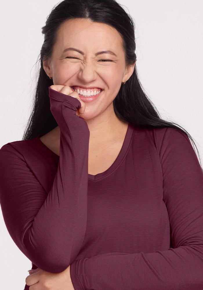 Smiling woman with long dark hair wears Woolx Millie Long Sleeve in Grape Harvest, hand by her mouth, eyes closed.