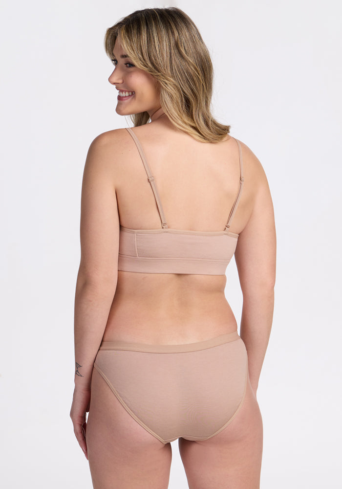 A woman with shoulder-length blonde hair, wearing the Woolx Luna Bralette - Morning Latte and matching underwear, stands smiling and facing away from the camera against a plain white background.