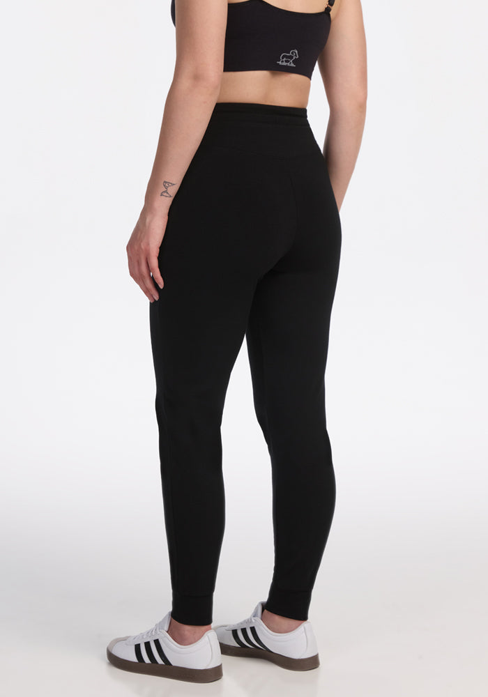 A person wearing a black sports bra, white sneakers with black stripes, and Woolx Madden Petite Joggers in black stands with their back turned against a plain white background.