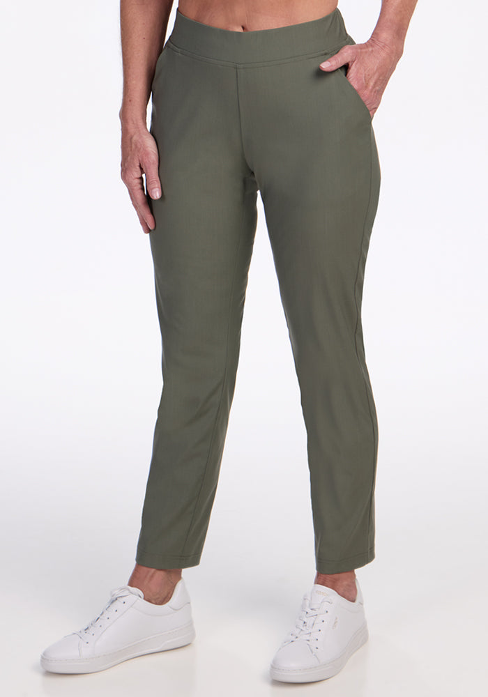 A person wearing Woolx Nicole Ankle Pants in Sage Olive and white sneakers stands with one hand in a pocket against a plain white background. 
