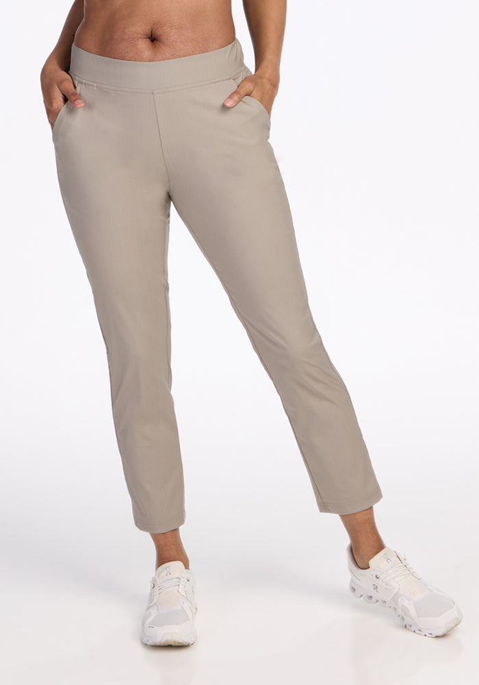 A person wears Woolx Nicole Ankle Pants in Vintage Khaki with white sneakers, standing hands-in-pockets against a plain white background. 