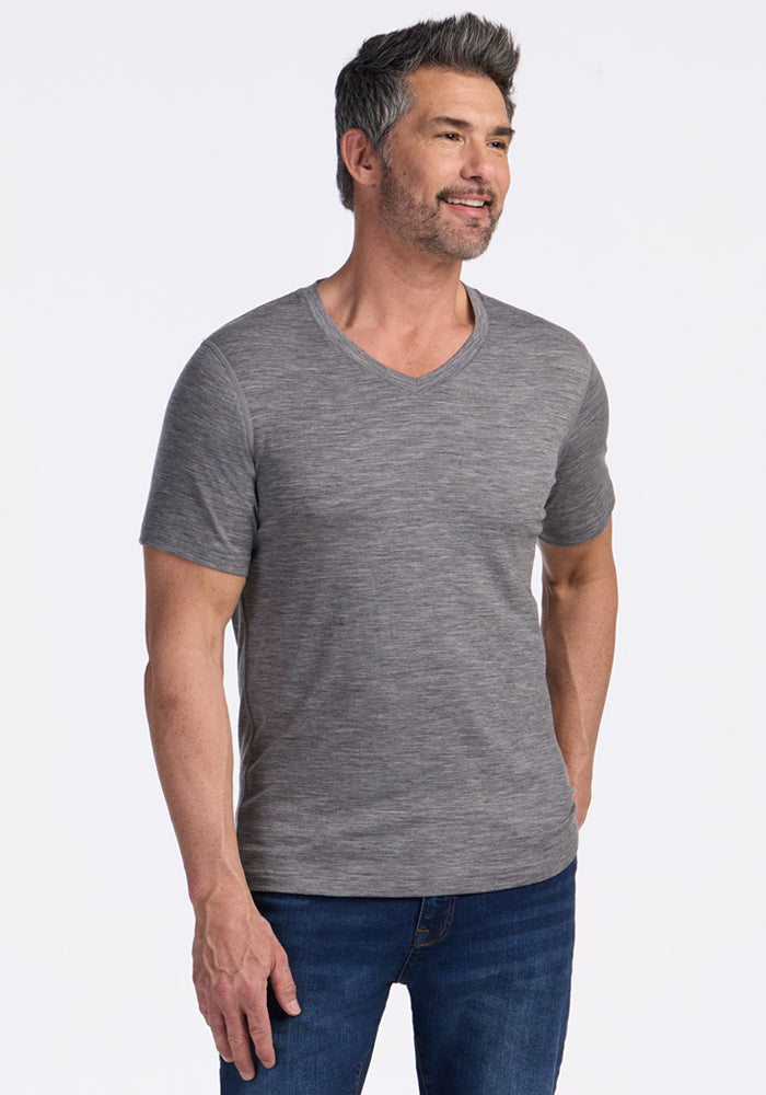A man with short dark and gray hair and a beard is smiling, wearing the Woolx Ashton V Neck in Graphite Heather and blue jeans, standing against a plain white background. 