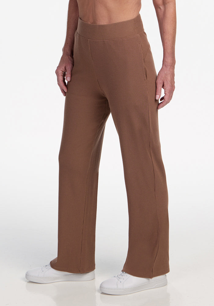 A person wearing Woolx Luca Ribbed Pants in Sienna Kiss with white sneakers stands against a plain white background. The image is cropped from the mid-torso down. 