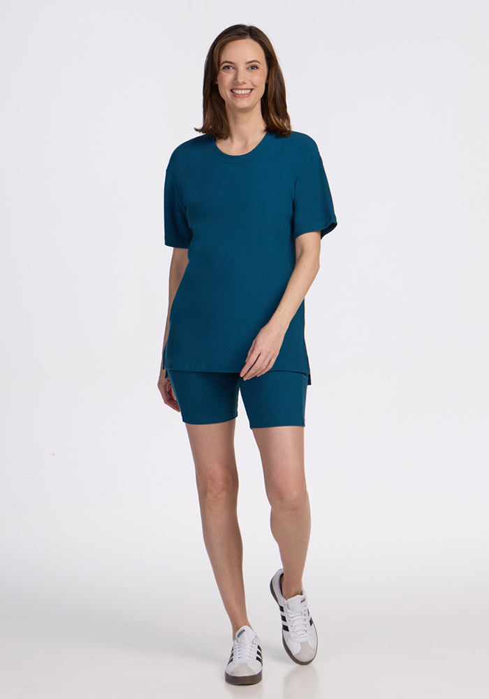 Woman smiling in the Woolx Vera Ribbed Tunic in Moroccan Blue, paired with matching shorts and white sneakers.