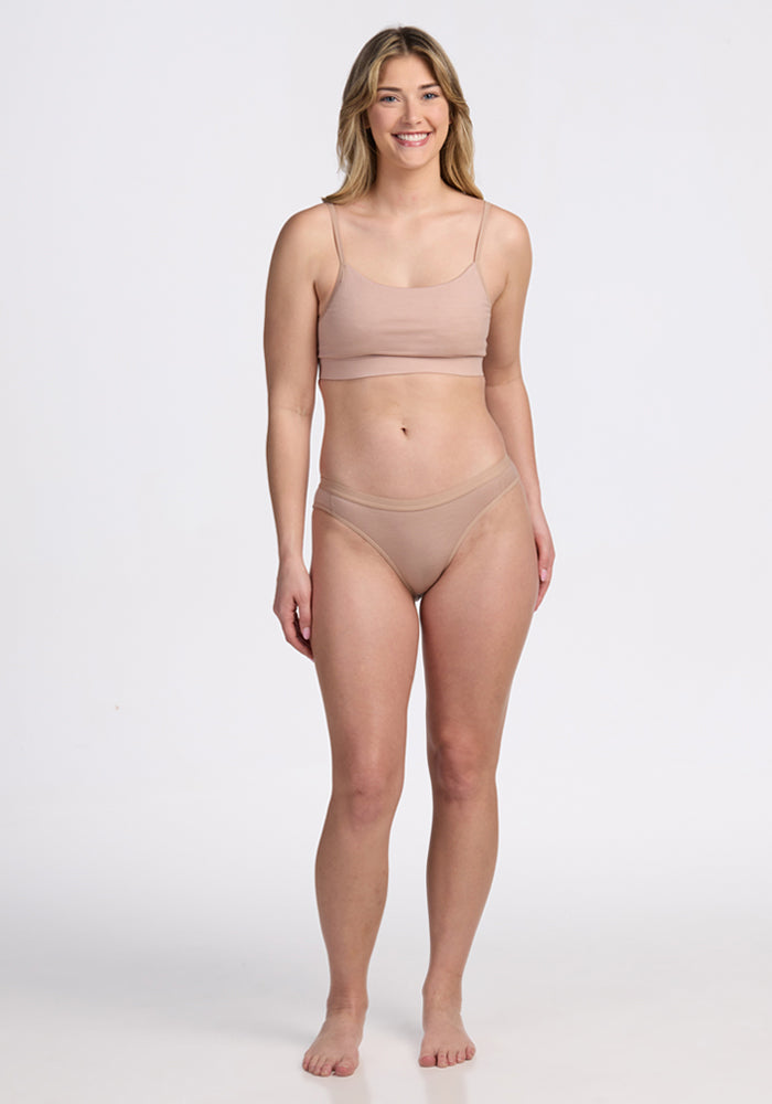 A woman with long, blonde hair is standing and smiling, wearing the Woolx Luna Bralette in Morning Latte and matching underwear against a plain white background.