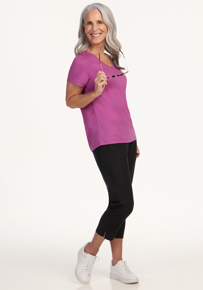 Smiling older woman in a Woolx Junie V Neck • Final Sale - Purple Wine, black pants, and white sneakers, holding eyeglasses.