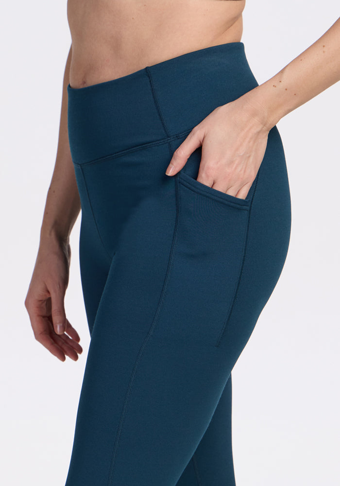 A person wears Woolx Merino Flex® McKenna Pocket Leggings in Majolica Blue, resting their right hand in the side pocket. The cropped image shows only the torso and upper legs against a plain background.