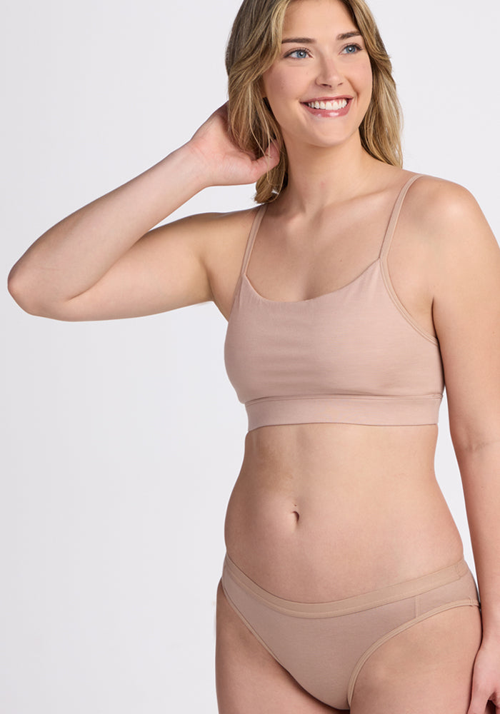 A woman with long, light brown hair smiles while wearing the Woolx Luna Bralette - Morning Latte and matching briefs, standing against a plain light background.