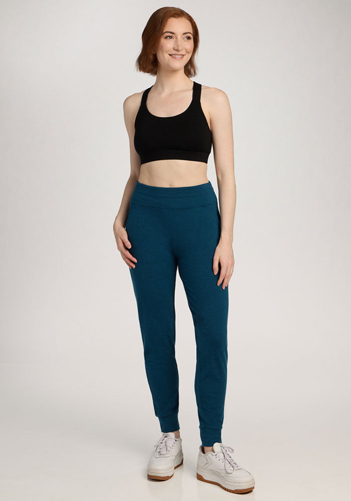 Woman smiling in Woolx Madden Petite Joggers in Majolica Blue, a black sports bra, and white sneakers.