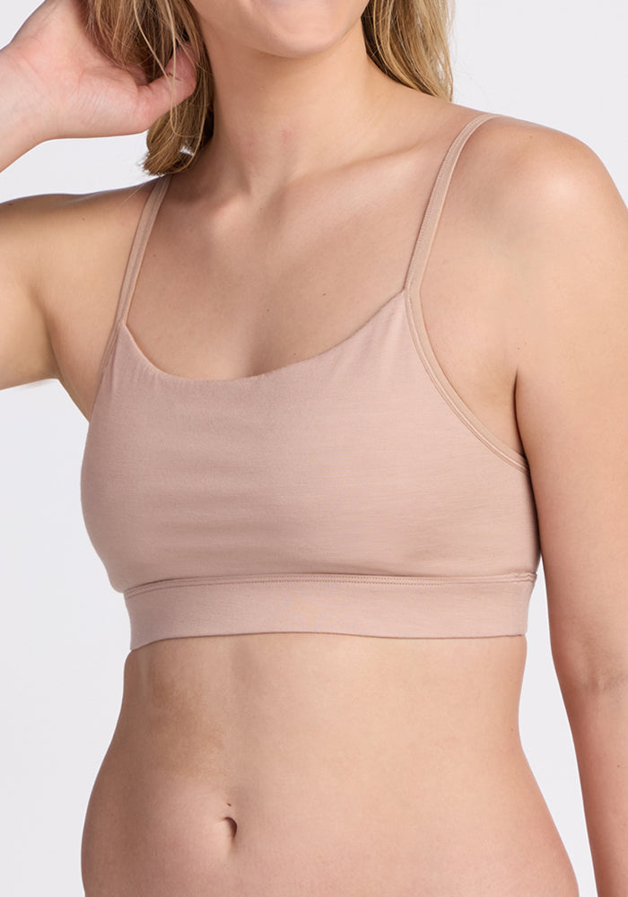 A woman wears the Woolx Luna Bralette in Morning Latte, a nude-hued, seamless, moisture-wicking bra with thin straps, standing against a plain background and touching her hair.