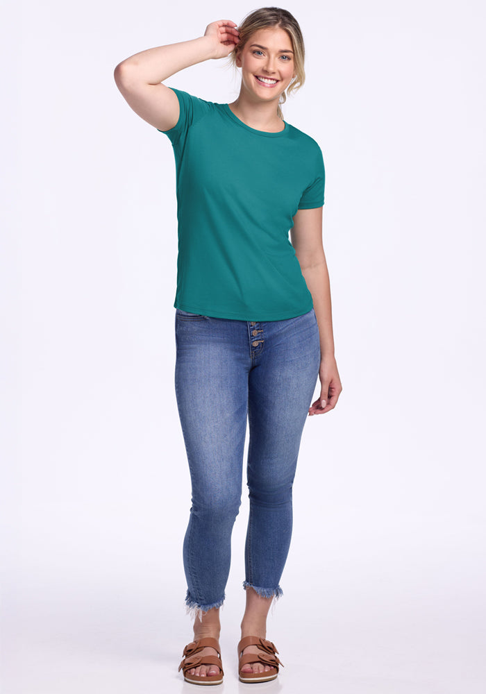 Smiling woman in the Woolx Liza Crew Neck Tee – Surfside, blue jeans, and sandals against a white background.
