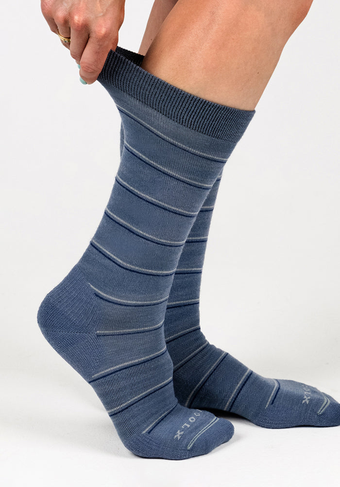 A person is pulling up a Woolx Womens Chandler Mid Calf Sock in Vintage Indigo, featuring light horizontal stripes. The lightweight, blue crew sock stands out against a plain white background.