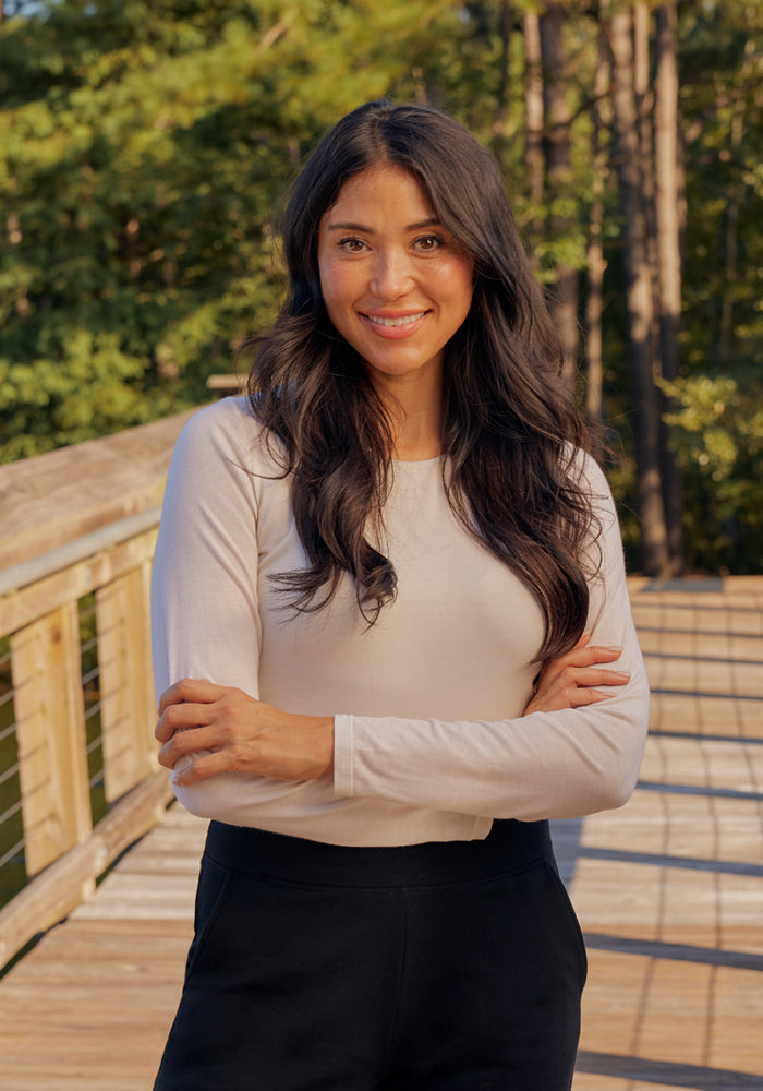 A woman with long dark hair, wearing the Woolx Sophia Bodysuit - Blue Horizon and black pants, stands outdoors on a wooden walkway, smiling with her arms crossed as sunlight filters through the trees in the inviting background.